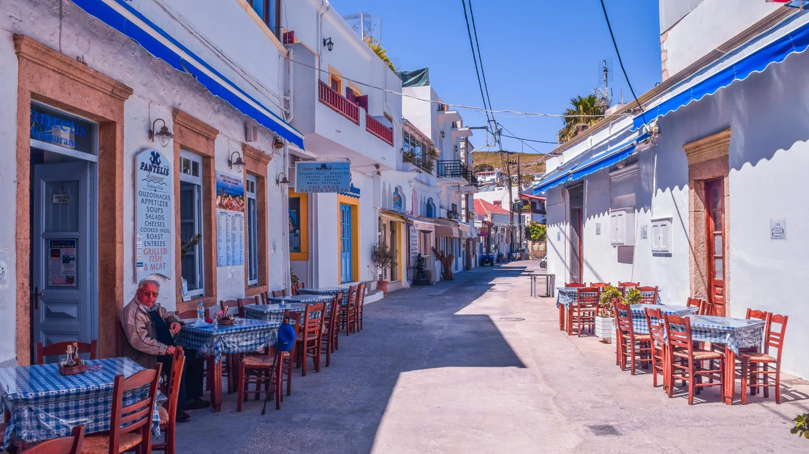 A picturesque Mediterranean street, tables and chairs in the sunshine. A lone old chap the only visible customer. 